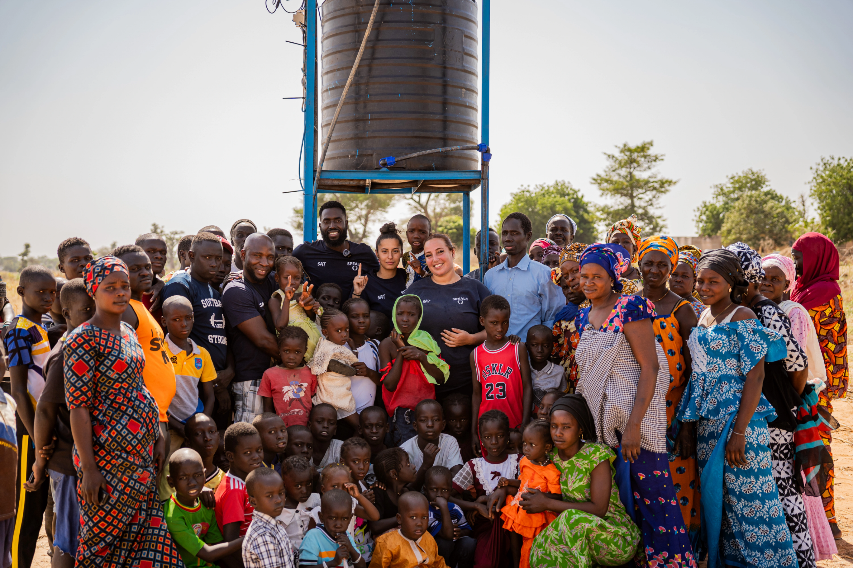 un grand groupe de personnes, incluant des hommes, femmes et enfants, pose ensemble devant un reservoir d'eau en afrique. Les visages souriants temoignent d'une ambiance festive