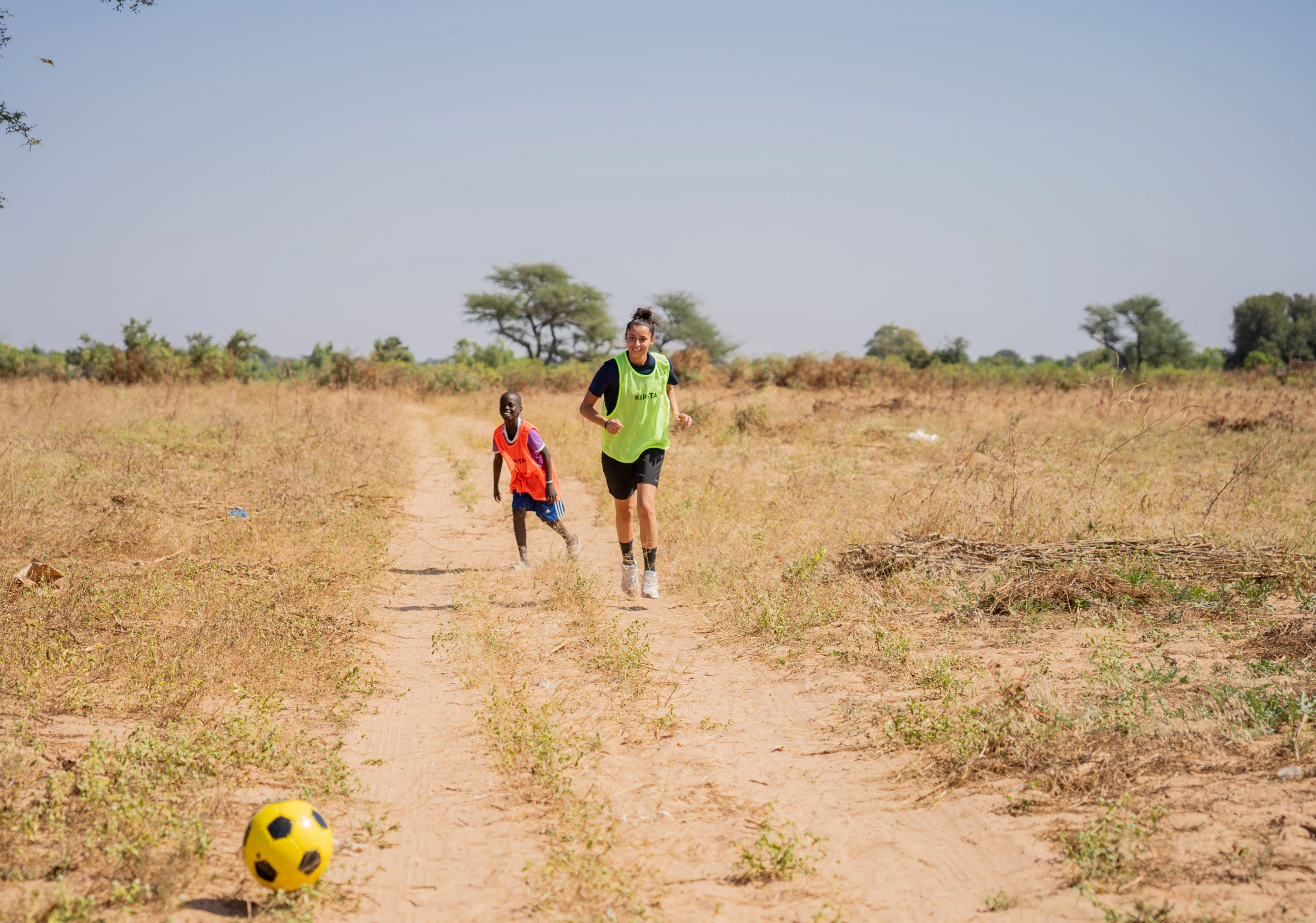 Deux jeunes jouant au football sur un terrain aride d'afrique courent après un ballon, l'un portant un maillot rouge et l'autre un maillot vert